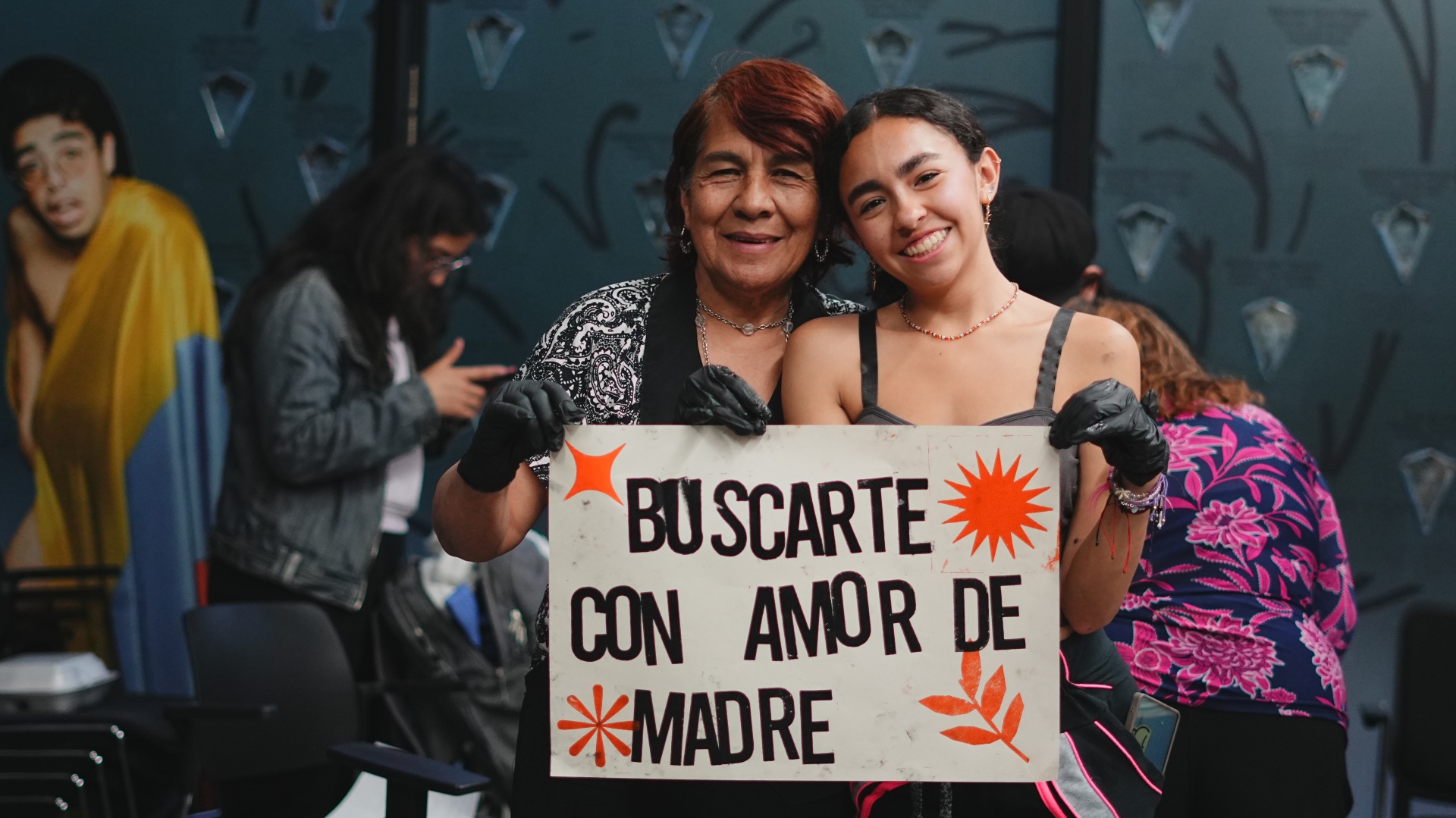 Mujer mayor con mujer joven, juntas sosteninendo cartel "Buscarte con amor de madre"